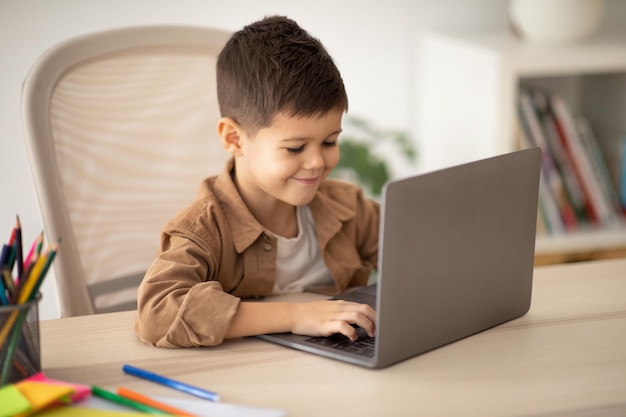 Videochiamata in remoto gioco educativo Felice bambino caucasico allievo si siede al tavolo digitando sul gioco al computer