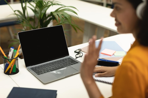 Videochiamata Insegnante nero che agita la mano sullo schermo vuoto del laptop che comunica online seduto al tavolo in biblioteca