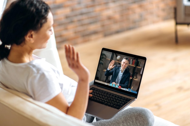 Videoconferenza di comunicazione online Una donna incinta seduta su un divano comunica in videochiamata con il suo amato marito o collega usando un laptop saluta l'interlocutore con un gesto della mano