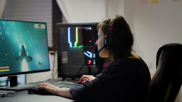 Videogioco professionale per donna r che utilizza cuffie e computer potente per giocare a videogiochi sparatutto spaziali online nella sala da gioco. Cibernetica in streaming virtuale che indossa le cuffie durante un torneo online