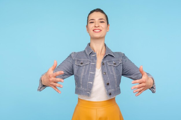 Vieni tra le mie braccia Ritratto di amichevole allegra donna vestita alla moda con crocchia per capelli che allunga le mani alla telecamera andando ad abbracciare dando un caloroso benvenuto girato in studio isolato su sfondo blu