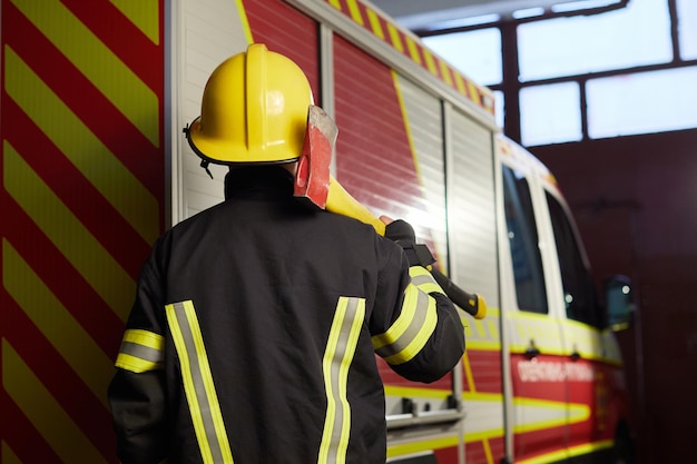 Vigile del fuoco completamente equipaggiato con casco e ascia sullo sfondo del camion dei pompieri Il soccorritore volta le spalle alla telecamera tenendo un'ascia in mano