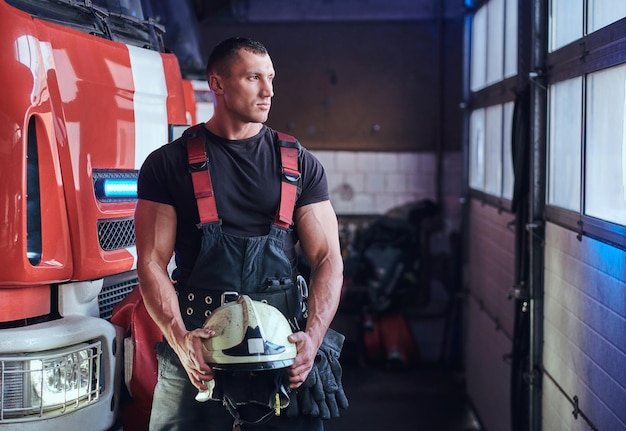 Vigile del fuoco muscolare che tiene un casco protettivo in un garage di un dipartimento dei vigili del fuoco, in piedi accanto a un'autopompa e guardando fuori