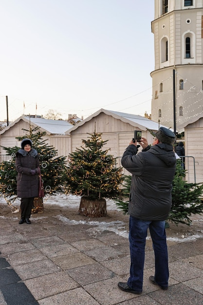 Vilnius, Lituania - 4 dicembre 2016: Persone che scattano foto al mercatino degli alberi di Natale con l'albero di Natale principale con ghirlande decorative sullo sfondo. Installato in Piazza della Cattedrale a Vilnius