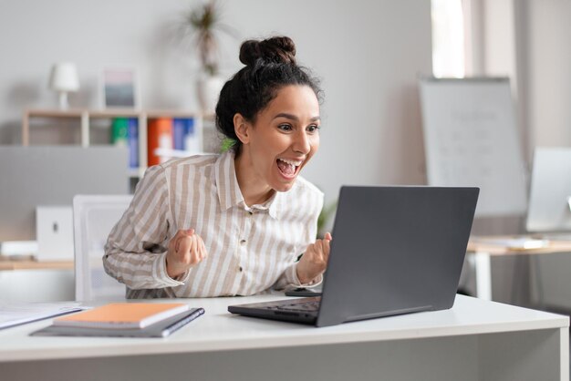 Vinci una donna d'affari eccitata emotiva che celebra il successo agitando i pugni chiusi e guardando il laptop