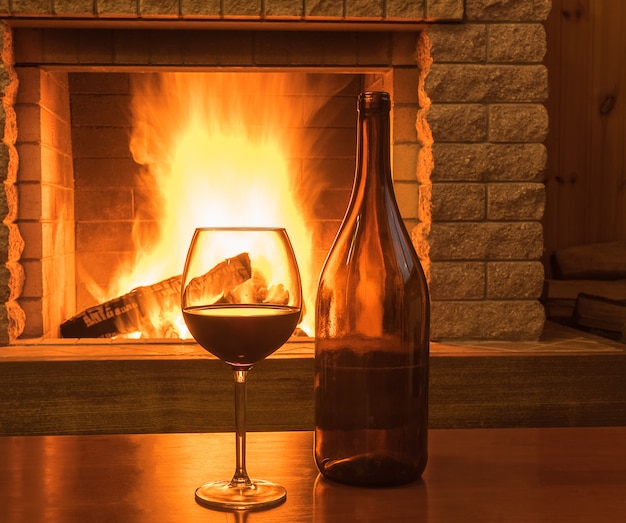 Vino rosso in un bicchiere e bottiglia, prima dell&#39;accogliente camino, in casa di campagna, inverno.