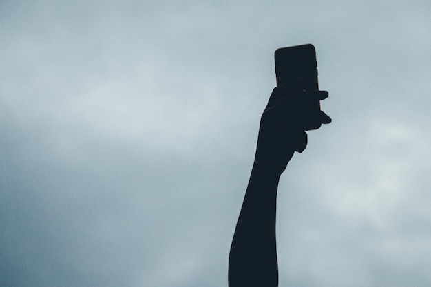 Vista a bassa angolazione della silhouette con la mano che tiene il telefono contro un cielo nuvoloso