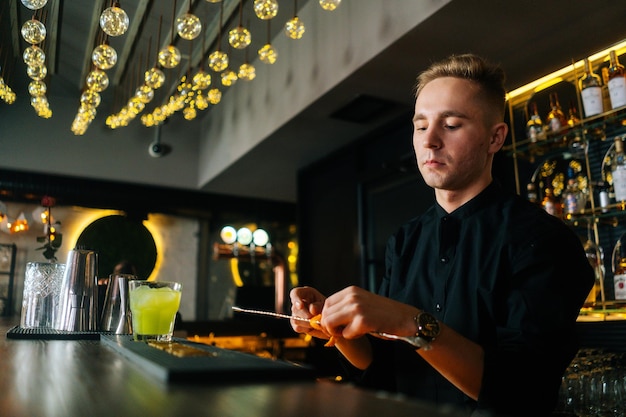 Vista a basso angolo del giovane barman barista maschio che spreme un pezzo di buccia d'arancia con paglia e decora un cocktail