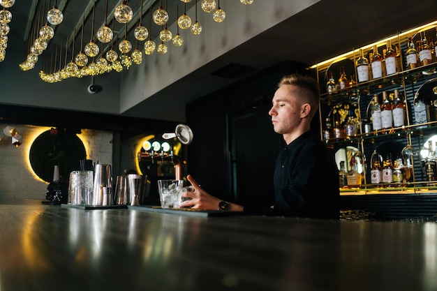 Vista a basso angolo di un barista serio che prepara cocktail alcolici rinfrescanti in piedi dietro il bancone del bar in una moderna discoteca buia, sullo sfondo di scaffali con diverse bevande alcoliche.