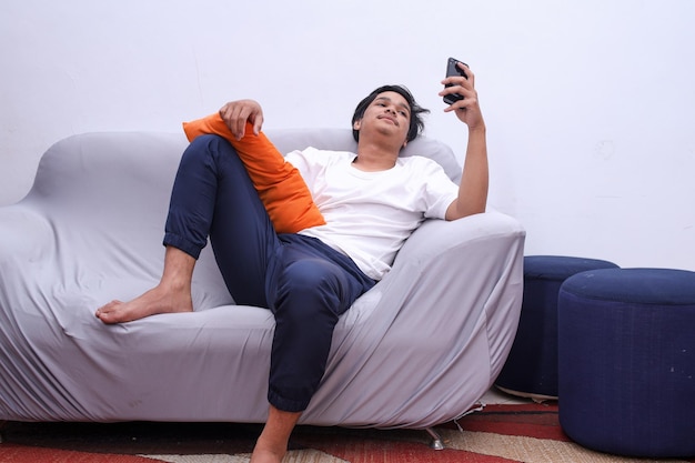 Vista a tutta lunghezza dell'uomo asiatico con smartphone sdraiato sul divano di casa Chat online sul telefono
