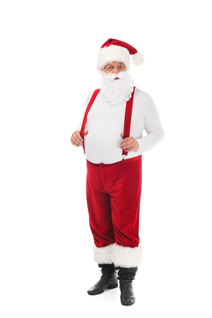 Vista a tutta lunghezza di Babbo Natale felice in cappello e bretelle che guarda l'obbiettivo isolato su bianco