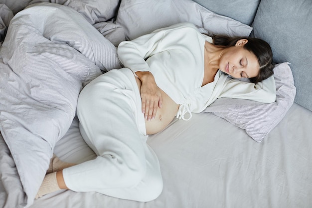 Vista ad alto angolo della giovane donna incinta in pigiama sdraiata nel suo letto durante il suo tempo libero