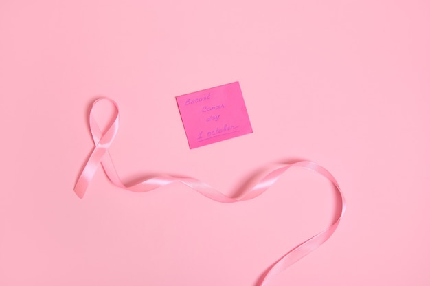 Vista ad alto angolo di un lungo nastro rosa con un'estremità infinita e una nota di carta con scritta 1 ottobre Consapevolezza del cancro al seno, isolata su sfondo rosa con spazio di copia. Lay piatto