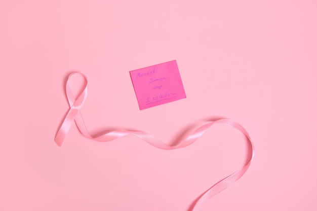 Vista ad alto angolo di un lungo nastro rosa con un'estremità infinita e una nota di carta con scritta 1 ottobre Consapevolezza del cancro al seno, isolata su sfondo rosa con spazio di copia. Lay piatto