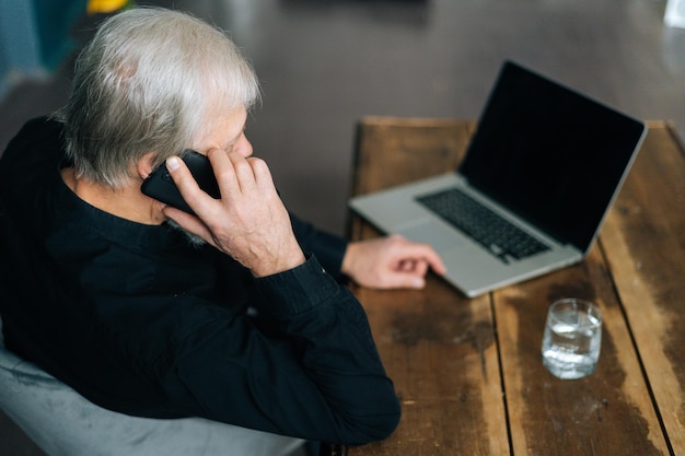 Vista ad alto angolo di un uomo adulto anziano dai capelli grigi irriconoscibile che parla al telefono cellulare seduto alla scrivania con un computer portatile. Primo piano del maschio più anziano maturo che ha conversazione sullo smartphone.