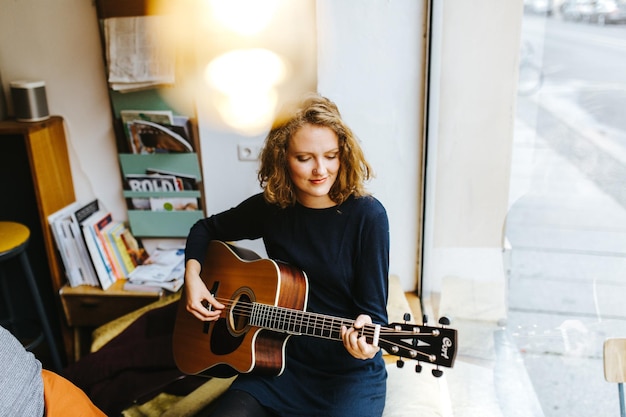 Vista ad alto angolo di una giovane donna che suona la chitarra davanti alla finestra di casa