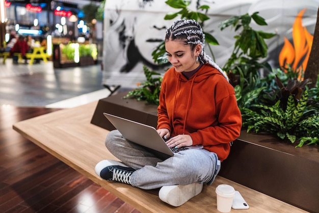 Vista ad alto angolo di una giovane donna che usa il suo laptop seduto su una panchina con le gambe incrociate in un centro commerciale