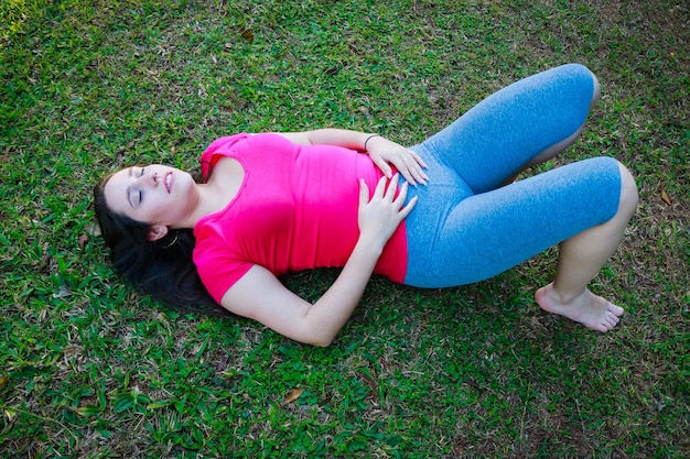 Vista ad alto angolo di una giovane donna con gli occhi chiusi e le mani sullo stomaco sdraiata sul campo erboso del parco