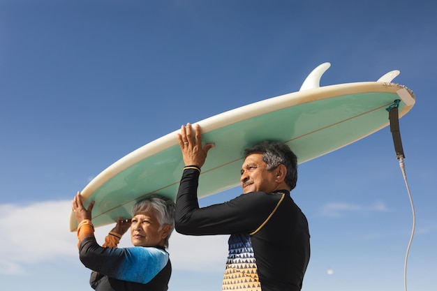 Vista ad angolo basso della coppia senior multirazziale che trasporta la tavola da surf sopra la testa contro il cielo blu