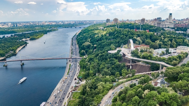 Vista aerea della nuova costruzione del ponte del parco ciclistico pedonale, fiume Dnepr, colline, parchi e paesaggio urbano di Kiev dall'alto, skyline della città di Kiev, Ucraina