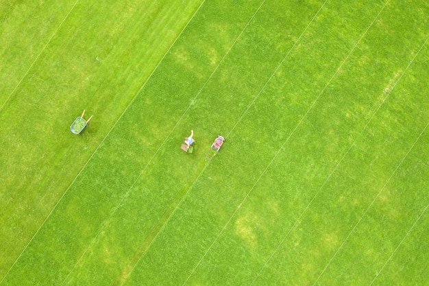 Vista aerea della piccola figura di un lavoratore uomo che taglia l'erba verde con la falciatrice sul campo dello stadio di calcio in estate.