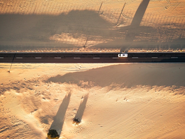 Vista aerea della strada asfaltata nera diritta con sabbia e deserto su entrambi i lati intorno - auto che viaggia nel mezzo - concetto di voglia di viaggiare per destinazioni esotiche e desertiche - luce e ombre sul