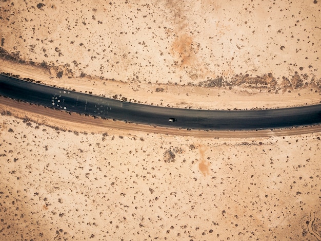 Vista aerea della strada asfaltata nera diritta con sabbia e deserto su entrambi i lati intorno - auto che viaggia nel mezzo - concetto di voglia di viaggiare per destinazioni esotiche e desertiche