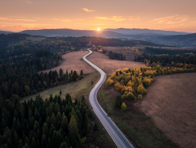 Vista aerea della strada di montagna nella foresta autunnale al tramonto in Ucraina Vista dall'alto dal drone della strada nei boschi Bellissimo paesaggio con l'autostrada nelle colline alberi prati cielo arancione luce solare dorata in autunno