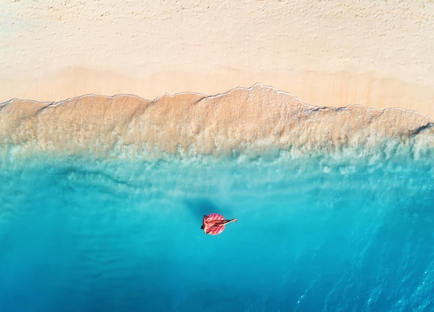 Vista aerea di una giovane donna che nuota con l'anello di nuoto a ciambella nel mare cristallino con onde al tramonto in estate Paesaggio aereo tropicale con ragazza acqua azzurra spiaggia sabbiosa Vista dall'alto Viaggio