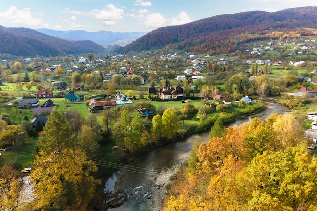 Vista aerea di una zona rurale del villaggio con piccole case tra colline di montagna autunnali ricoperte di foreste di abete rosso giallo e verde.