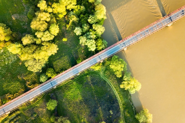 Vista aerea di uno stretto ponte stradale che si estende su un ampio fiume fangoso nella verde zona rurale.