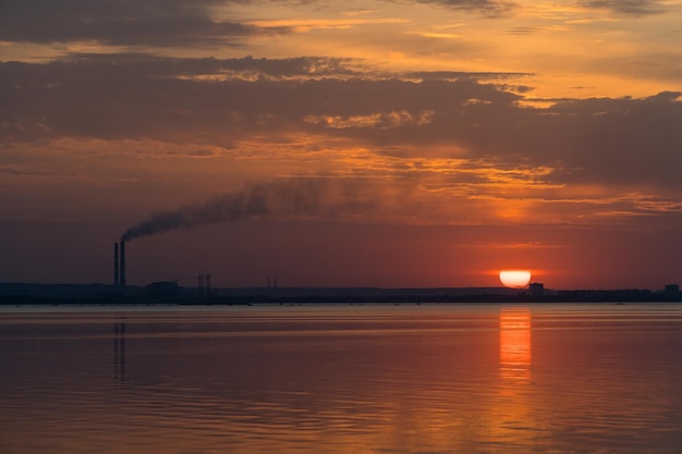 Vista al tramonto di tubi industriali fumatori su un orizzonte sopra il fiume