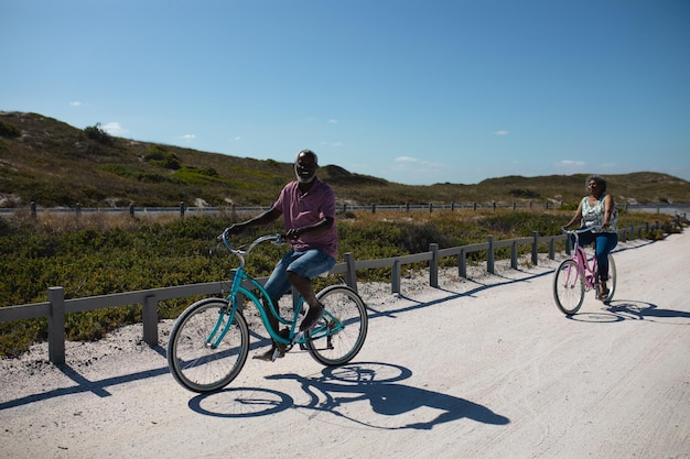Vista anteriore di una coppia di anziani afroamericani che guidano le biciclette sulla spiaggia con cielo blu e altopiani sullo sfondo