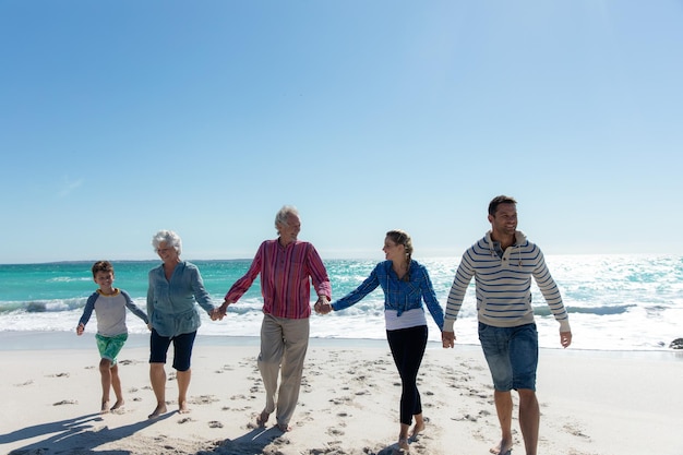 Vista anteriore di una famiglia caucasica multigenerazionale sulla spiaggia con il cielo blu e il mare sullo sfondo, che cammina, si tiene per mano e sorride