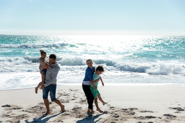 Vista anteriore di una famiglia caucasica sulla spiaggia con il cielo blu e il mare sullo sfondo, che si abbracciano e si divertono