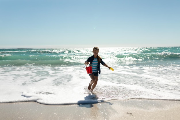 Vista anteriore di una ragazza caucasica su una spiaggia soleggiata, che corre a piedi nudi sulla sabbia verso la telecamera con un secchio e una pala, con il cielo blu e il mare sullo sfondo