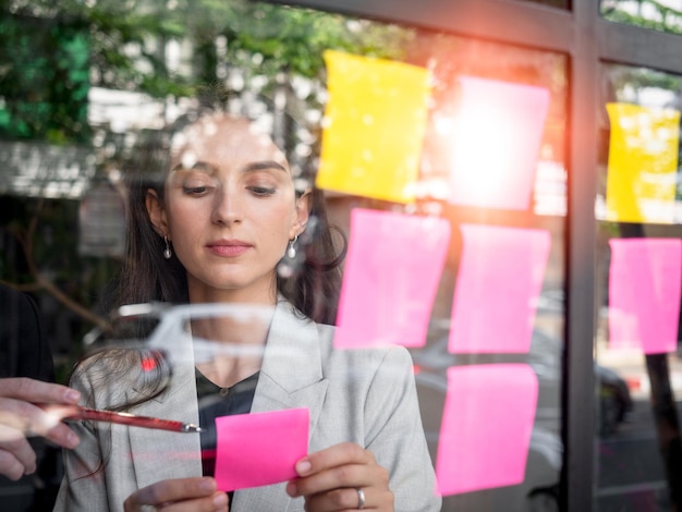 Vista attraverso il vetro imprenditrice che lavora alla strategia con un collega allegato postit colorati sulla parete di vetro dove idee scritte piano d'azione per il raggiungimento dell'obiettivoxA