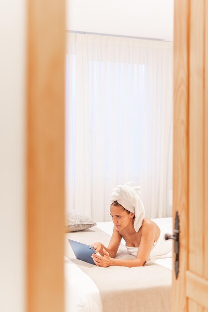 Vista attraverso la porta di una donna avvolta in asciugamani che usa il suo tablet digitale mentre è sdraiata sul letto della camera da letto di un hotel. Concetto di vacanza, hotel.