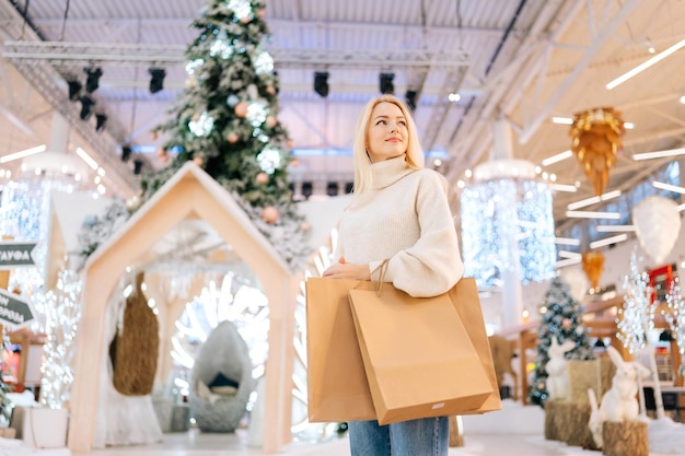 Vista dal basso del ritratto di giovane donna bionda carina che tiene borse con acquisto in piedi nella sala del centro commerciale celebra la vigilia di Natale