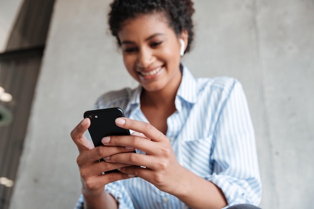 Vista dal basso di una giovane donna africana sorridente che indossa una maglietta che ascolta musica con auricolari wireless mentre è seduta al muro di casa e usa il telefono cellulare