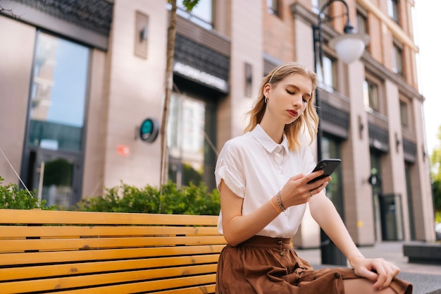 Vista dal basso di una giovane donna concentrata con lunghi capelli biondi in abiti casual che utilizza lo smartphone seduto su una panchina sulla strada della città sullo sfondo dell'edificio