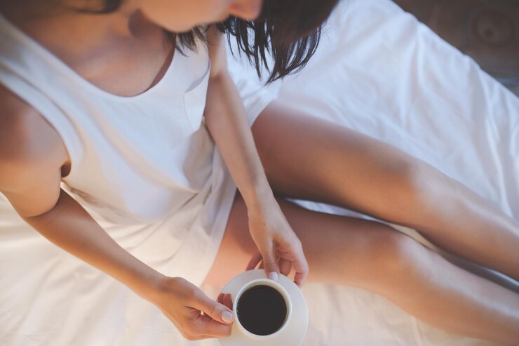 Vista dall'alto bella donna rilassante nell'accogliente atmosfera di casa sul letto bianco con una tazza di cioccolata calda o caffè in mano godendosi il comfort Sfondo chiaro soffuso Gambe attraenti