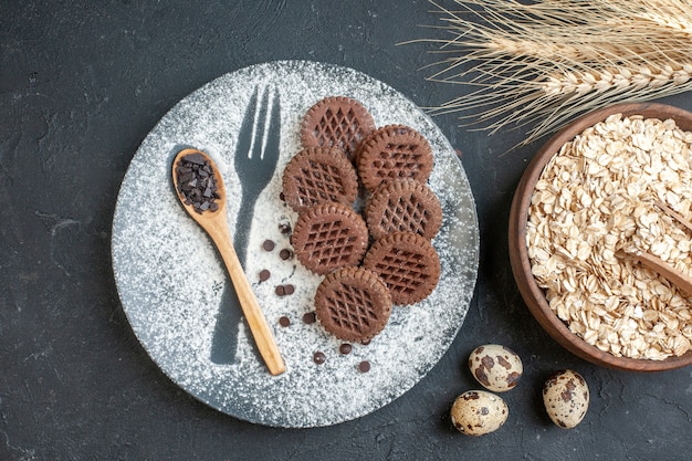 vista dall'alto biscotti al cacao cucchiaio di legno su piastra con forchetta impronta con zucchero a velo avena in una ciotola spighe di grano uova di quaglia su sfondo scuro