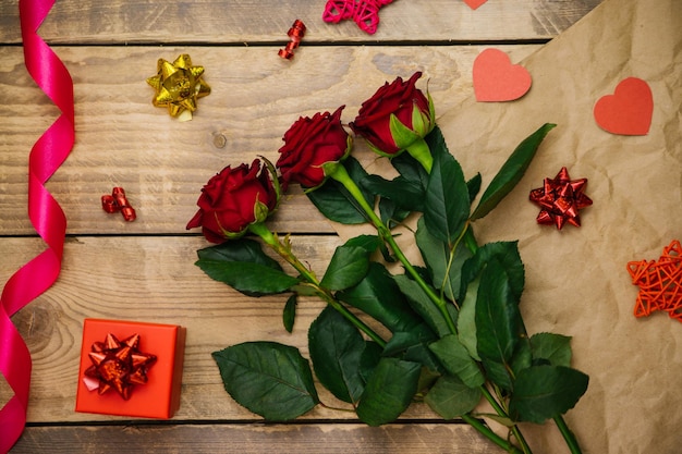 Vista dall'alto, confezione regalo rossa. Atmosfera festosa, bouquet di rose rosse, nastro di raso e fiocchi su fondo in legno.