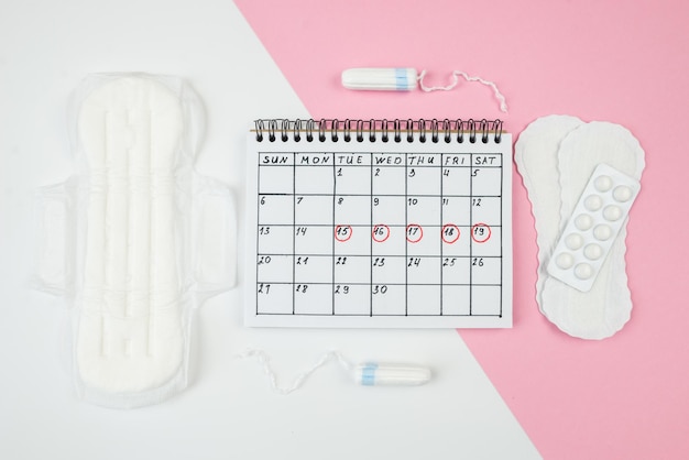 Vista dall'alto del calendario dei giorni critici delle donne con assorbenti e accessori su sfondo bianco e rosa Il concetto di mestruazioni femminili