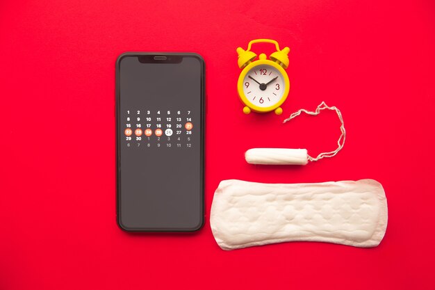 Vista dall'alto del calendario delle mestruazioni in smartphone con tampone di cotone, assorbente e sveglia.
