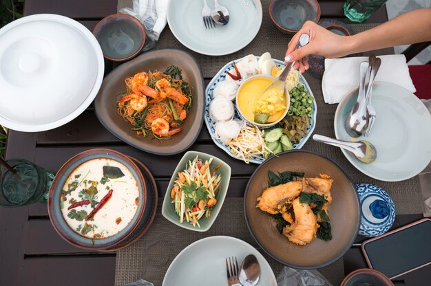 Vista dall'alto del cibo tailandese con granchio salato, spaghetti di riso, branzino fritto, mango affettato piccante, zuppa di pollo cremosa, insalata di vermicelli e stoviglie sul tavolo del ristorante