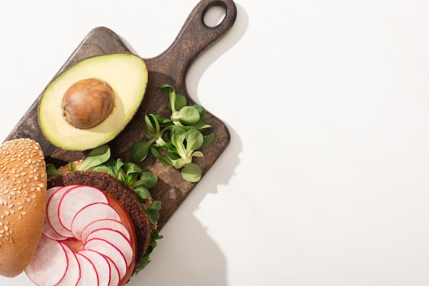 Vista dall'alto del delizioso hamburger vegano con ravanello avocado e verdure sul tagliere di legno su bianco