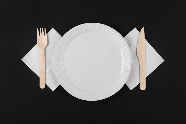 Vista dall'alto del piatto di carta bianca, forchetta di legno e coltello su tovagliolo bianco su sfondo nero