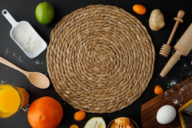 Vista dall'alto del sottopentola con uovo limone lime arancia mandarino farina di kumquat zenzero succo d'arancia con cucchiaio miele mestolo e mattarello su sfondo nero